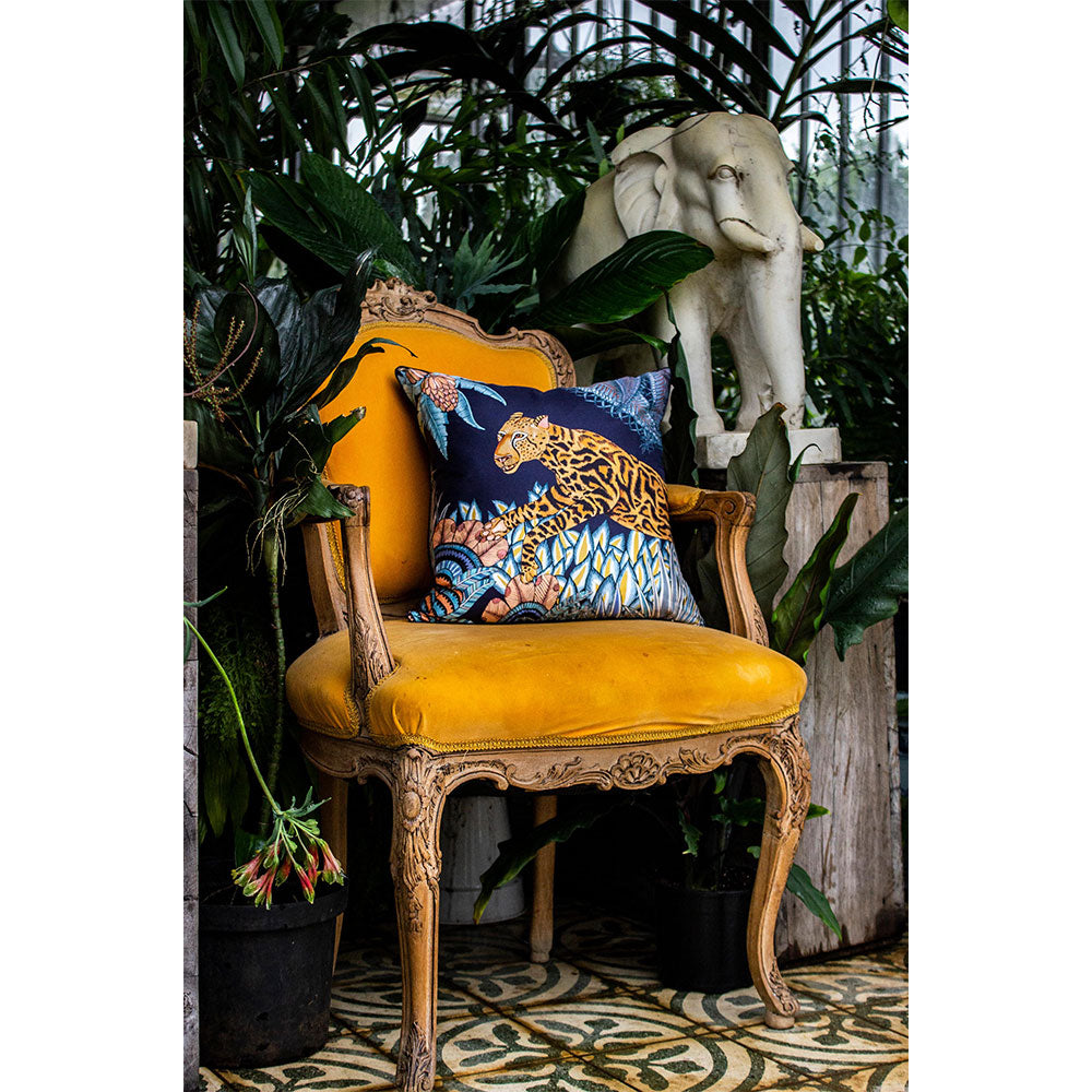 Lifestyle view of the navy Cheetah Kings Forest Pillow styled on a vintage mustard yellow velvet armchair set in a lush greenhouse environment alongside an elephant statue.