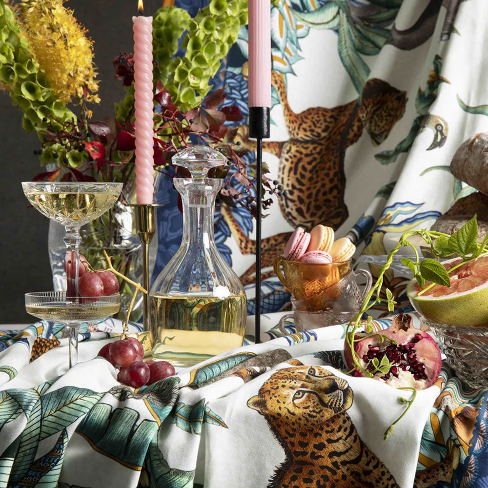 Ardmore wildlife tablecloth styled on a dining table with leopard motifs, tropical greenery, pink candles, glassware, fruit, and coordinating linens.