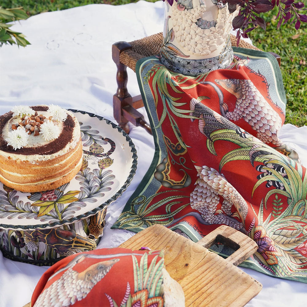 Red Ardmore pangolin-print tea towel styled at an outdoor picnic with coordinating linens, wooden serving boards, and a layered cake.