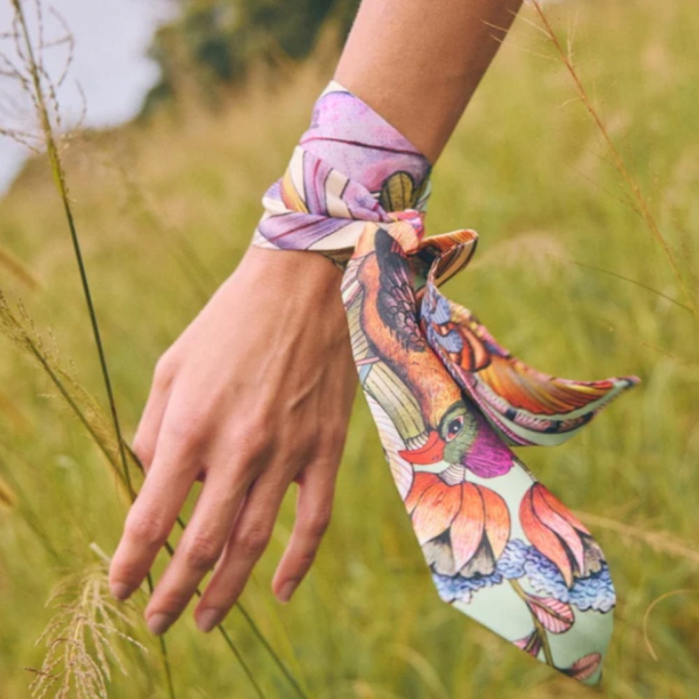 Close-up of a model wearing the Phinda Pride Flamingo Silk Twill scarf wrapped around their wrist like a bracelet, set against a blurry grassy field outdoors.