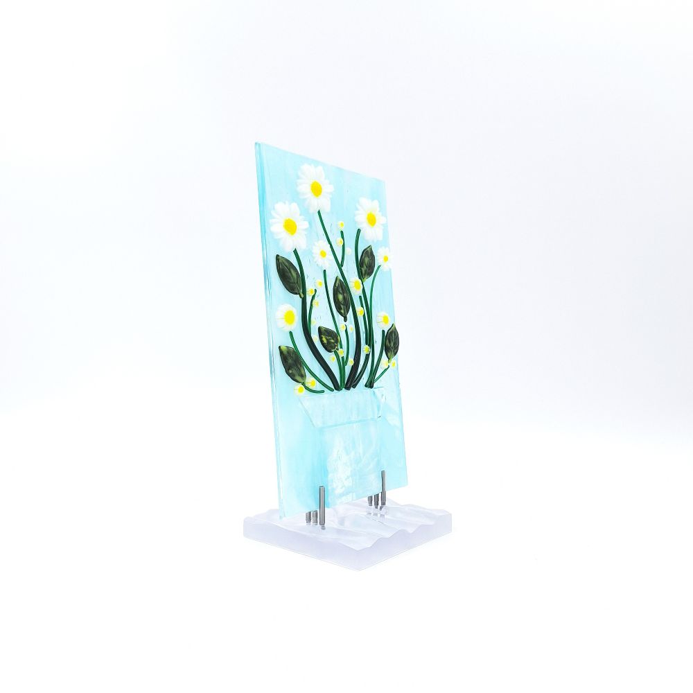 Angled view of the White Daisies Panel featuring white flowers with yellow centers, green stems, and dark green leaves on a pale blue glass background with a textured white base.