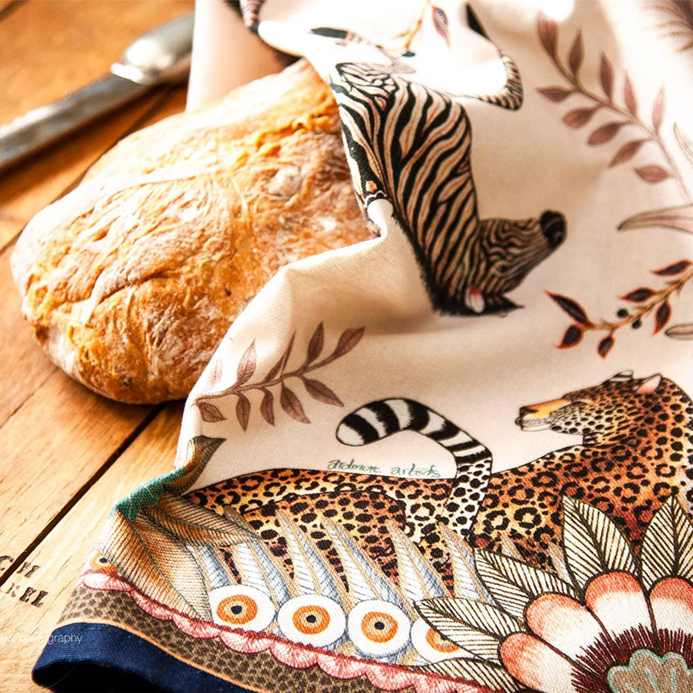 Bush Bandits Tea Towel styled on a wooden surface beside a loaf of bread, showing zebra and leopard artwork with botanical details and a decorative border.