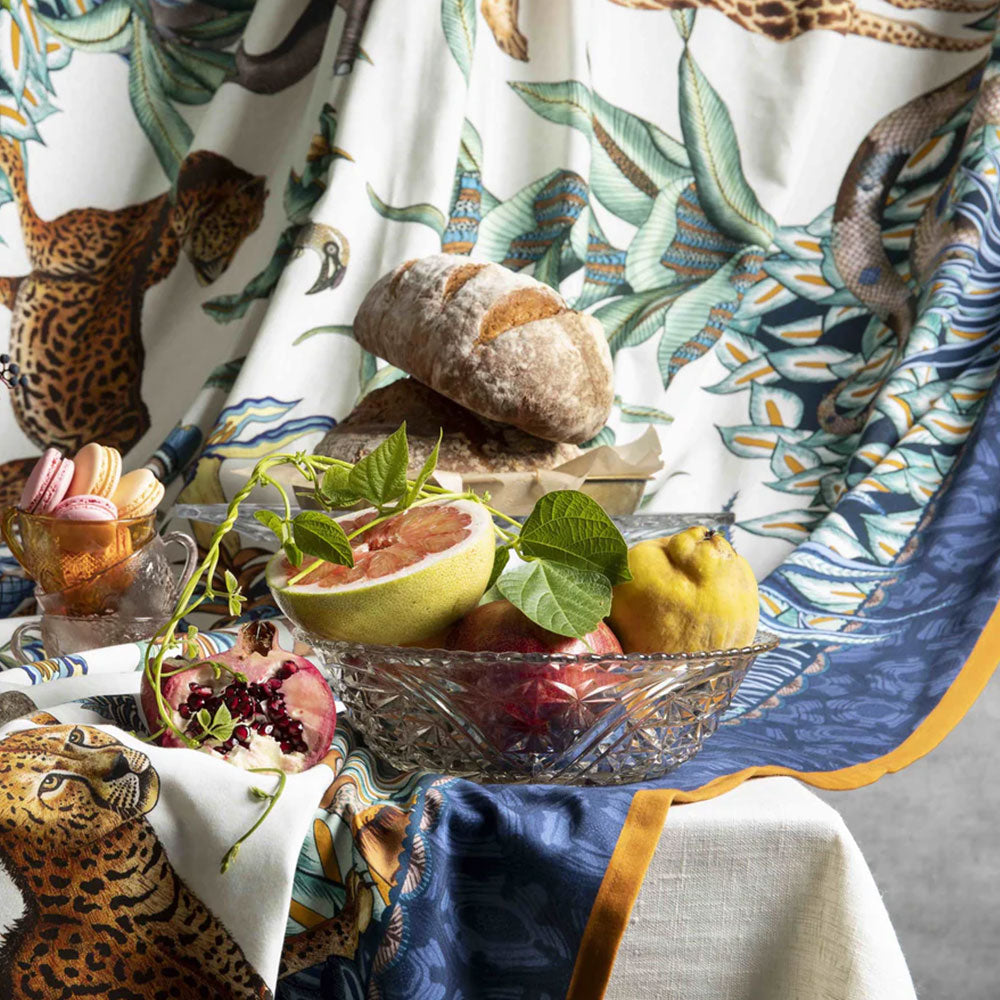 Still life arrangement on a table style with the Sabie Forest tablecloth, featuring a glass bowl filled with fresh fruit including a halved grapefruit, pomegranate, and quince, flanked by artisanal bread and a small dish of pastel macarons.