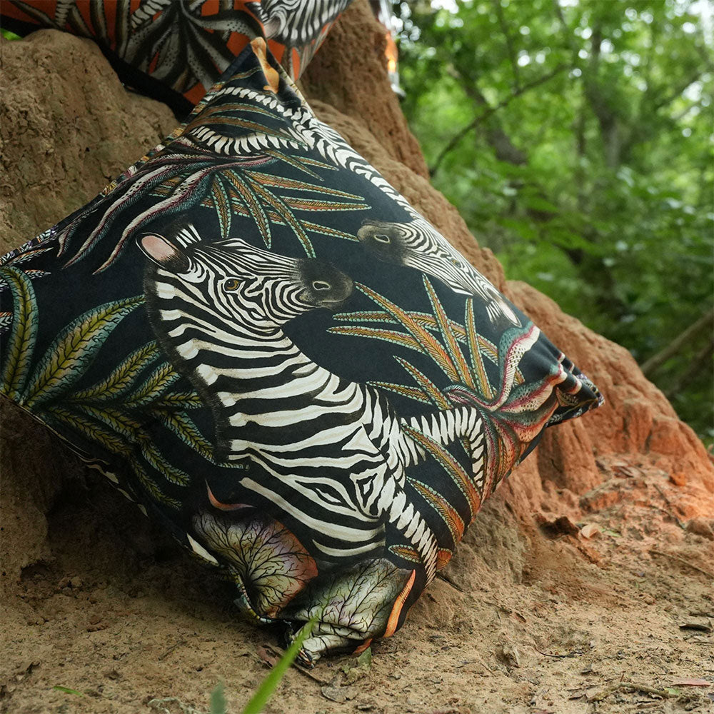 Thanda Stripe Pillow styled outdoors against a tree, featuring illustrated zebras surrounded by detailed botanical foliage in cream, olive, gold, and earthy tones on a black background.