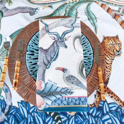 Ardmore wildlife-print tea towel styled on a layered place setting with a kudu, bird, leopard, tropical foliage, and coordinating table linens.