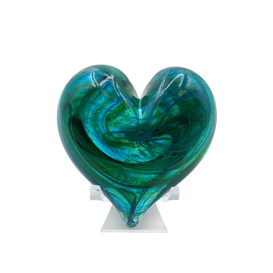 Handmade heart-shaped glass sculpture featuring swirling teal, turquoise, emerald green, and deep blue tones, displayed on a clear stand against a white background.