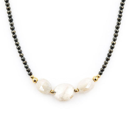 Close-up of a centered necklace featuring faceted dark pyrite beads, three pale peach moonstone beads, and polished gold-tone accents on a white background.