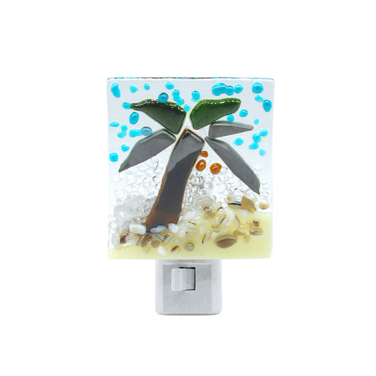 Front view of a handmade fused glass nightlight featuring a tropical palm tree scene with turquoise blue accents, green palm leaves, and textured sandy beige glass on a white plug-in base.