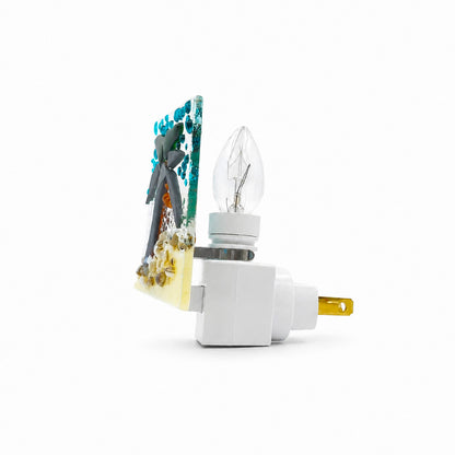 Side view of a handmade tropical fused glass nightlight showing the textured palm tree glass panel, clear bulb, white plug-in base, and gold-tone prong.