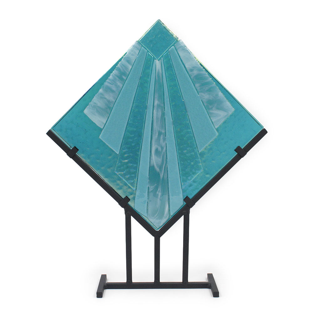 Abstract turquoise glass sculpture with layered geometric panels arranged in a diamond shape, displayed on a black metal stand against a white background.