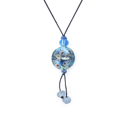 Close-up of a black cord necklace featuring a round aqua-blue glass pendant with raised silver-tone and multicolored details, blue accent beads, and two dangling pale blue glass beads.