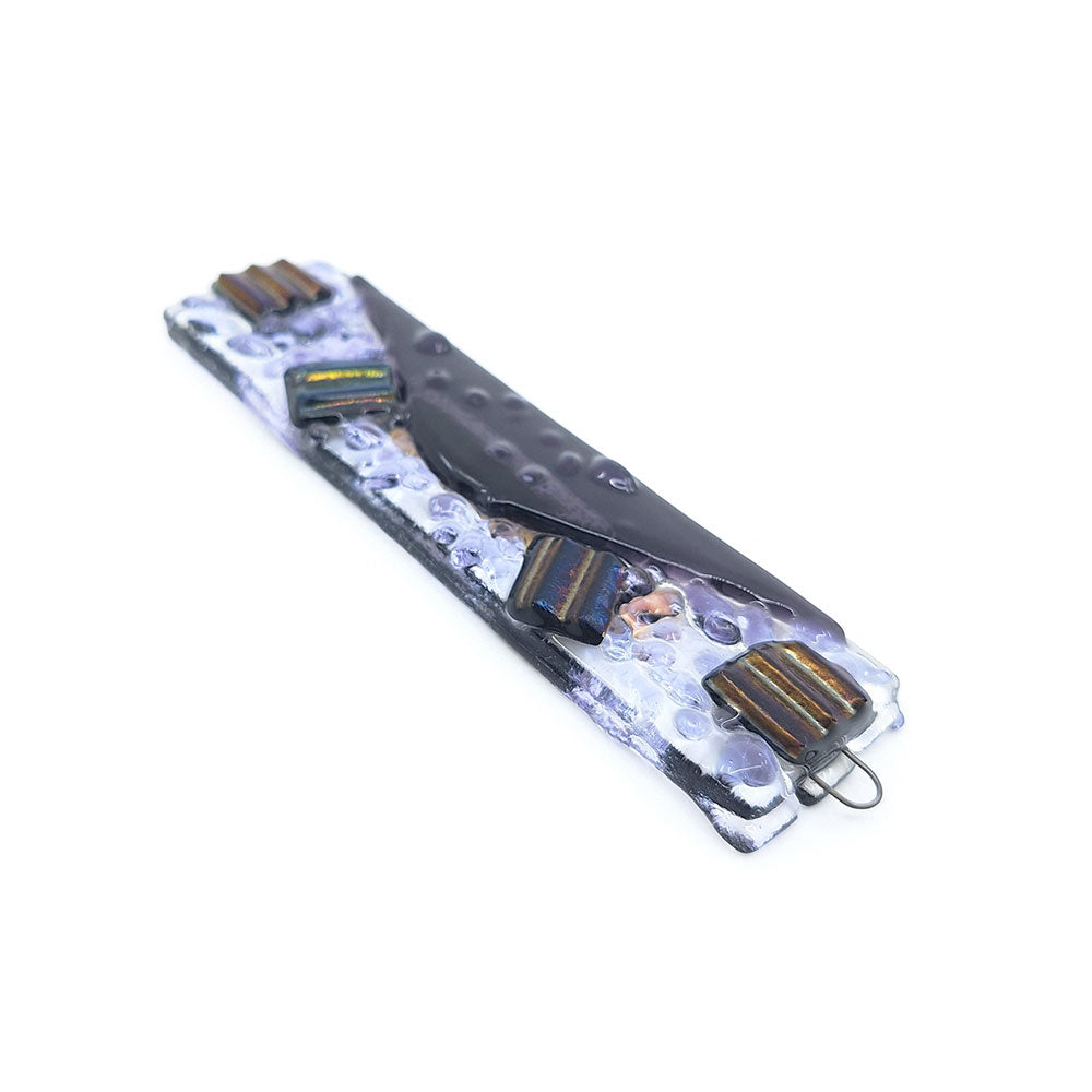 Handmade glass mezuzah featuring a clear textured rectangular base with a dark sculptural center, iridescent striped glass accents, and metal hanging loops on a white background.