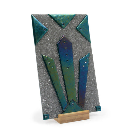 Angled view of an abstract glass sculpture with a textured silver background, layered iridescent teal and green geometric glass pieces, metallic accents, and a light wood display base.