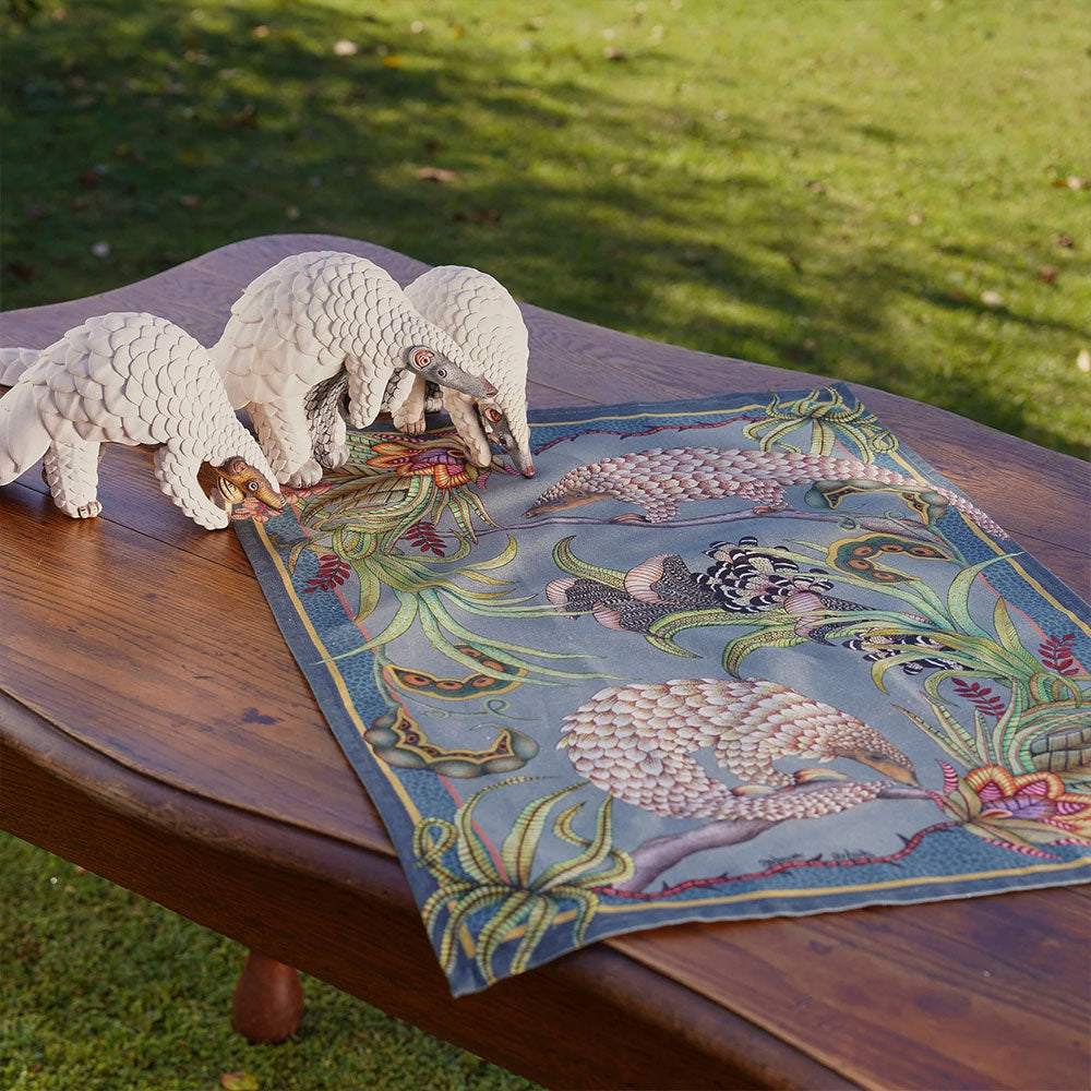 Ardmore pangolin-print tea towel displayed on a wooden outdoor table beside three decorative pangolin figurines.