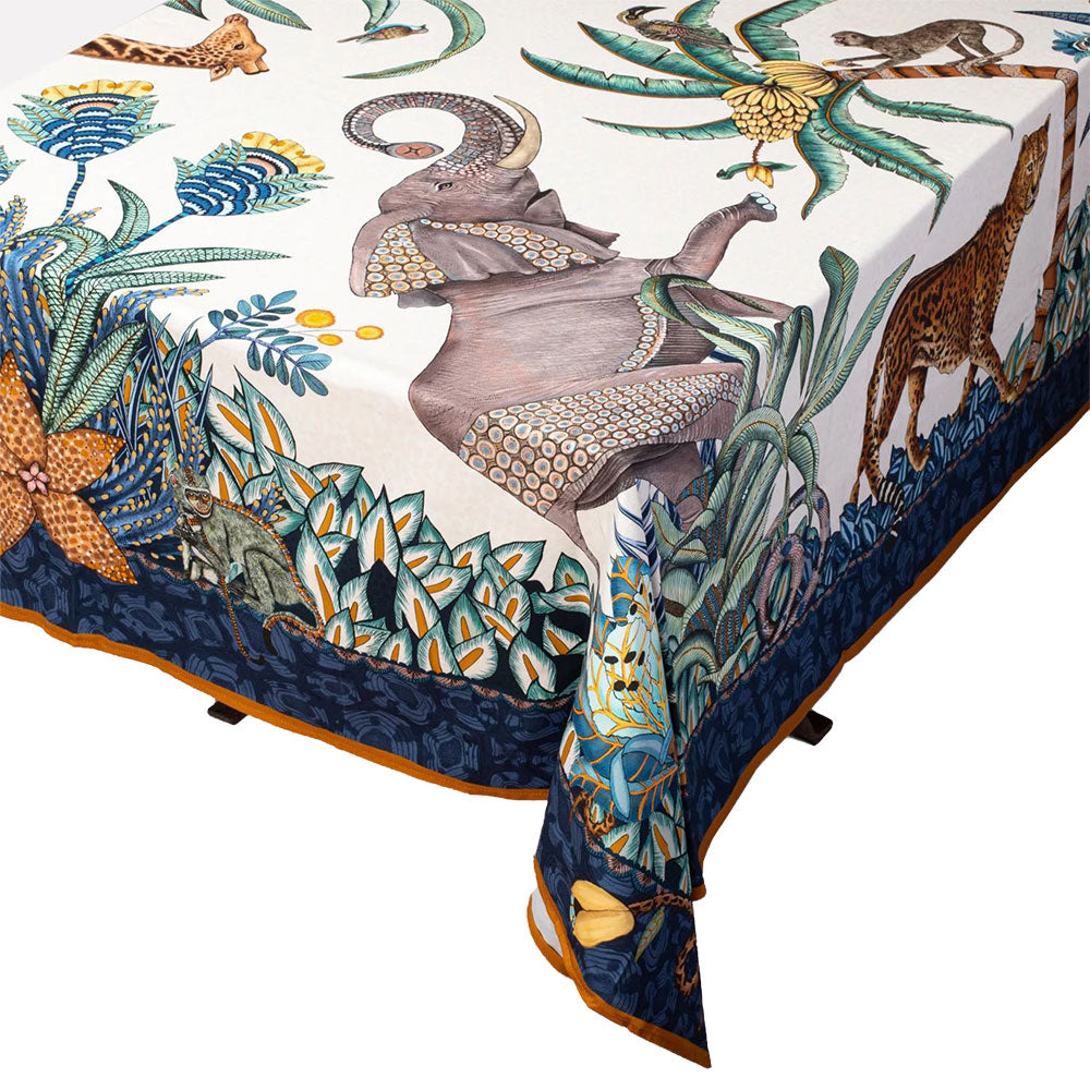 Close-up of an Ardmore wildlife tablecloth draped over a table, showing elephants, a giraffe, tropical plants, and an ornate blue border.