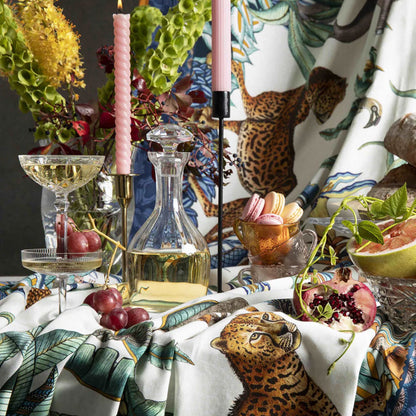 Ardmore wildlife tablecloth styled on a dining table with leopard motifs, tropical greenery, pink candles, glassware, fruit, and coordinating linens.