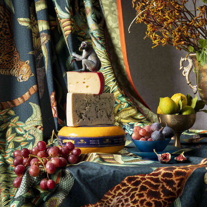Deep green Ardmore wildlife tablecloth styled with layered fabric, stacked cheeses, grapes, figs, pears, and decorative animal accents.