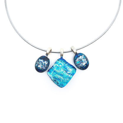 Close-up of a stainless steel necklace featuring three blue dichroic glass charms suspended from sterling silver pinch bails, with a large square center charm flanked by two smaller oval charms.
