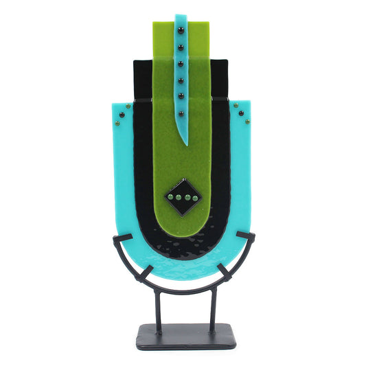 Abstract freestanding glass sculpture in turquoise, lime green, and black with layered geometric panels, metallic stud accents, and a dark metal display stand on a white background.