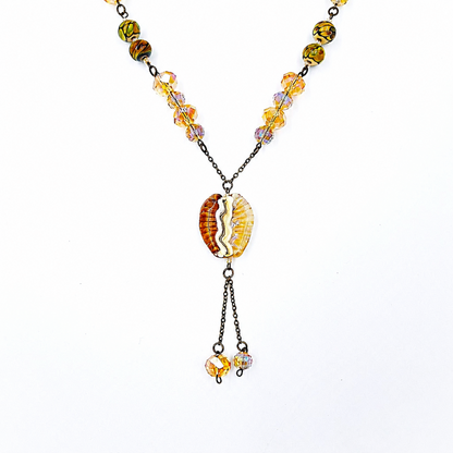 Close-up of a handmade amber lariat necklace showing faceted golden glass beads, patterned green and amber beads, an amber and cream focal bead, antique-bronze chain, and coordinating dangling bead drops.