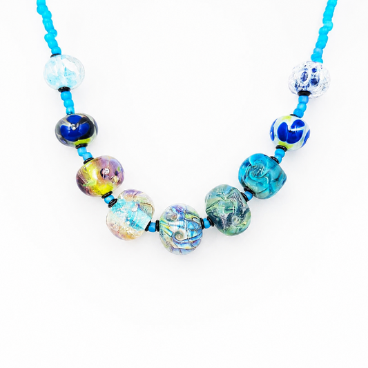 Close-up of a turquoise beaded necklace featuring assorted decorative glass beads in blue, aqua, green, purple, and gold tones on a white background.