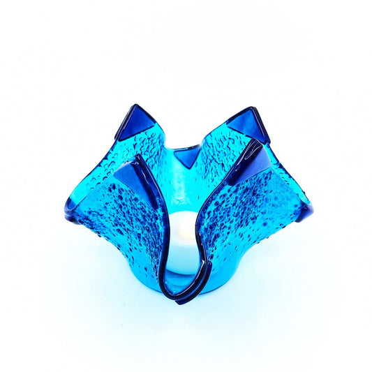Angled view of a handmade blue glass tulip candle holder with curved asymmetrical petals, textured aqua glass, and dark blue edging on a white background.