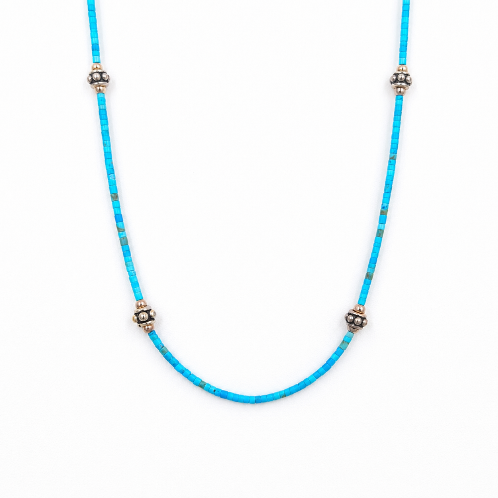 Close-up of a straightened turquoise beaded necklace featuring tiny rectangular beads and four evenly spaced silver-tone floral accents on a white background.