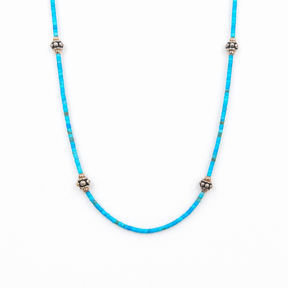 Close-up of a straightened turquoise beaded necklace featuring tiny rectangular beads and four evenly spaced silver-tone floral accents on a white background.