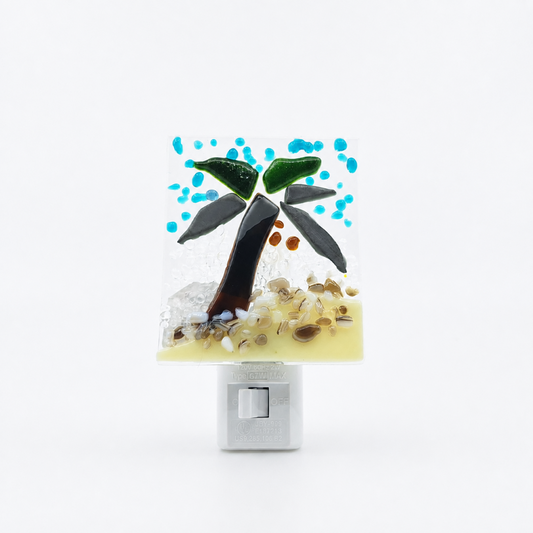 Front view of a handmade fused glass nightlight featuring a tropical palm tree scene with turquoise blue accents, green palm leaves, and textured sandy beige glass on a white plug-in base.