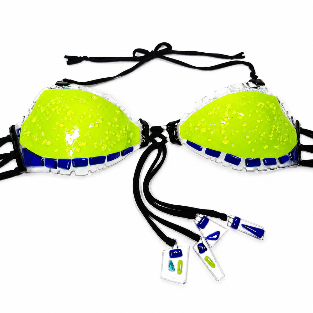 Close-up of a handmade glass bikini top showing textured neon lime green glass cups with cobalt blue and clear glass accents, black ties, and small decorative glass charms.