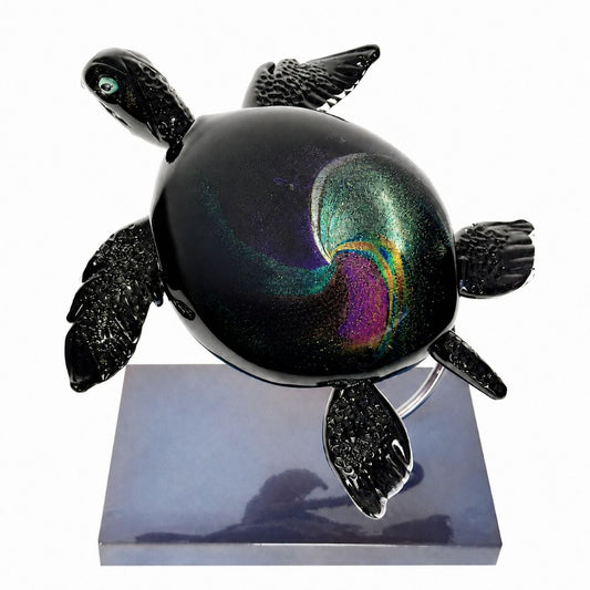 Iridescent black glass sea turtle sculpture with textured flippers and a swirling galaxy-inspired shell in teal, green, purple, pink, and gold, displayed on a dark rectangular stand.