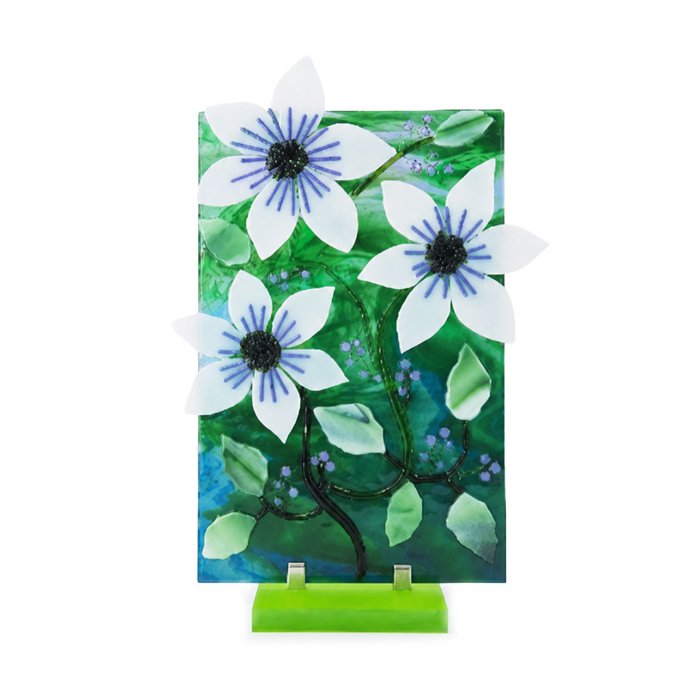 Front view of a decorative floral glass panel featuring three dimensional white flowers with blue centers, green leaves, dark winding stems, and small blue and purple accents on a bright green stand.