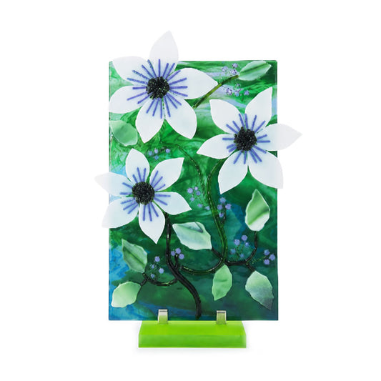 Front view of a decorative floral glass panel featuring three dimensional white flowers with blue centers, green leaves, dark winding stems, and small blue and purple accents on a bright green stand.