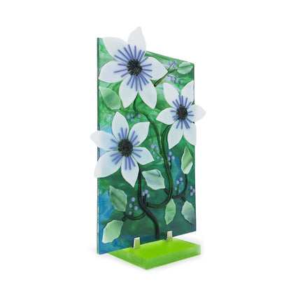 Angled view of a decorative floral glass panel featuring raised white flowers with blue centers, layered green leaves, dark curving stems, and blue and purple accents displayed on a bright green stand.