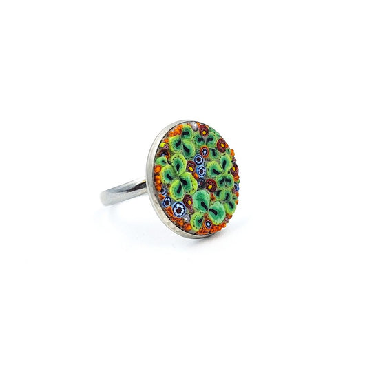 Silver-tone circular ring featuring a colorful mosaic glass face with green floral shapes and orange, red, blue, black, and white accents, displayed on a white background.