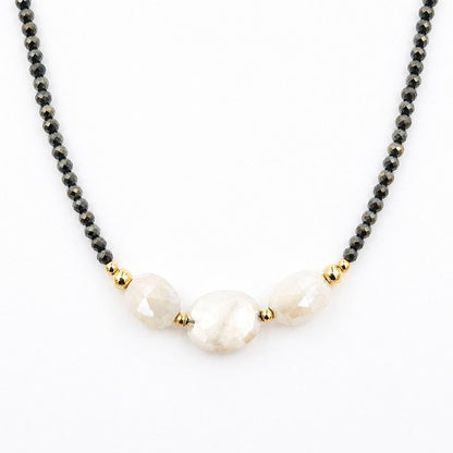 Close-up of a centered necklace featuring faceted dark pyrite beads, three pale peach moonstone beads, and polished gold-tone accents on a white background.