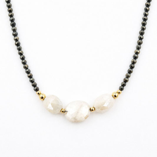 Close-up of a centered necklace featuring faceted dark pyrite beads, three pale peach moonstone beads, and polished gold-tone accents on a white background.