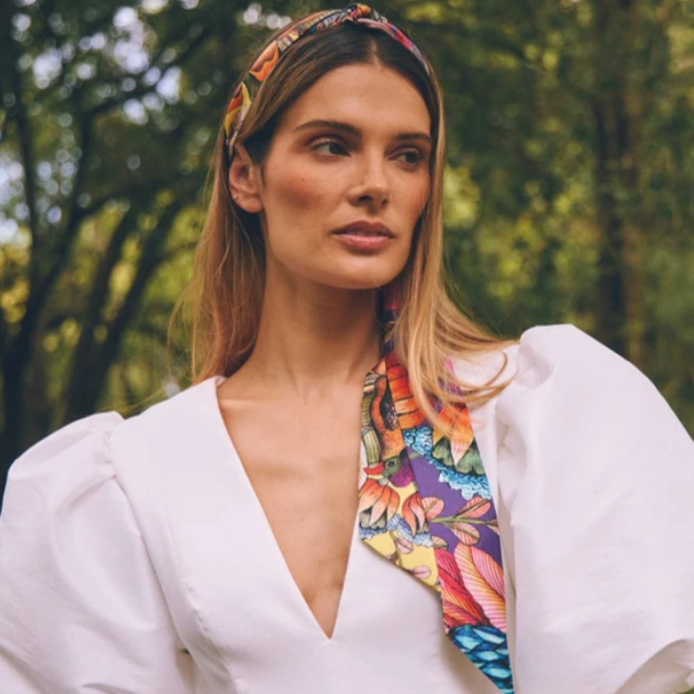 Model wearing the Phinda Pride Paradise Silk Twill scarf as a headband with long ends draping over her shoulder, styled with a white puff-sleeve top outdoors. 