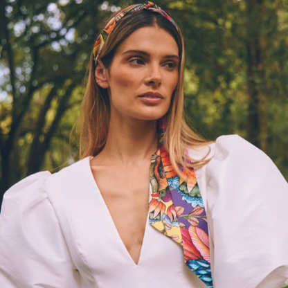 Model wearing the Phinda Pride Paradise Silk Twill scarf as a headband with long ends draping over her shoulder, styled with a white puff-sleeve top outdoors. 