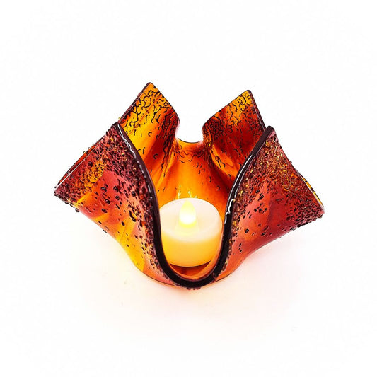 Handmade red and amber glass tulip candle holder shown with a glowing candle inside, featuring sculpted asymmetrical petals, textured glass, and deep red edging.