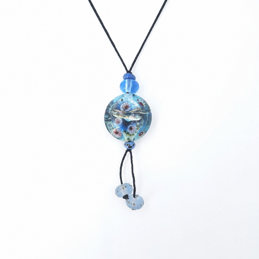 Close-up of a black cord necklace featuring a round aqua-blue glass pendant with raised silver-tone and multicolored details, blue accent beads, and two dangling pale blue glass beads.