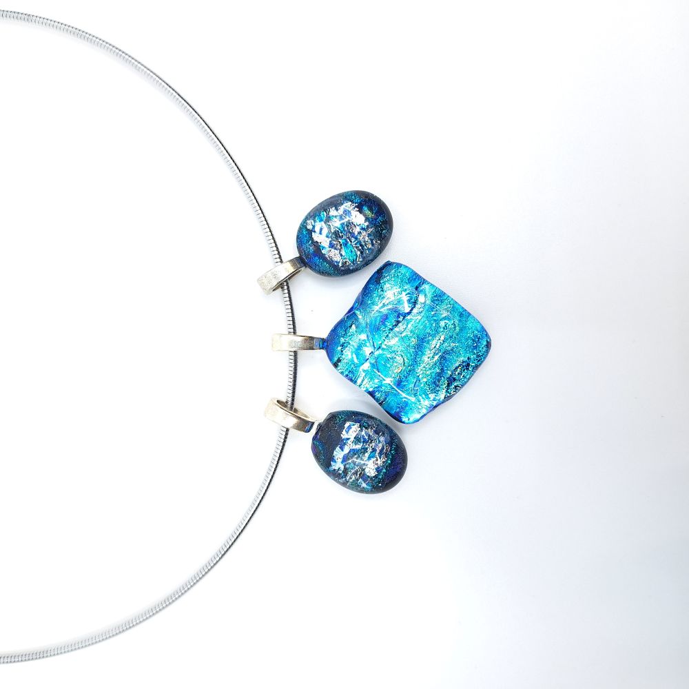 Close-up of a stainless steel necklace featuring three blue dichroic glass charms suspended from sterling silver pinch bails, with a large square center charm flanked by two smaller oval charms.