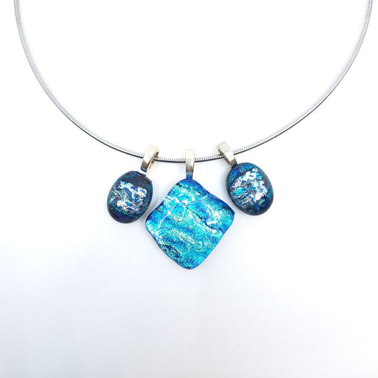 Close-up of a stainless steel necklace featuring three blue dichroic glass charms suspended from sterling silver pinch bails, with a large square center charm flanked by two smaller oval charms.