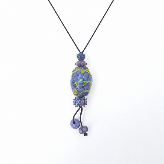 Handmade pendant necklace featuring a black cord, layered purple and blue lampwork glass beads, floral and vine details, and two dangling lavender bead accents on a white background.
