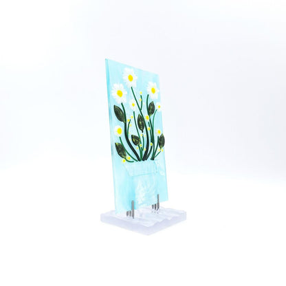 Angled view of the White Daisies Panel featuring white flowers with yellow centers, green stems, and dark green leaves on a pale blue glass background with a textured white base.