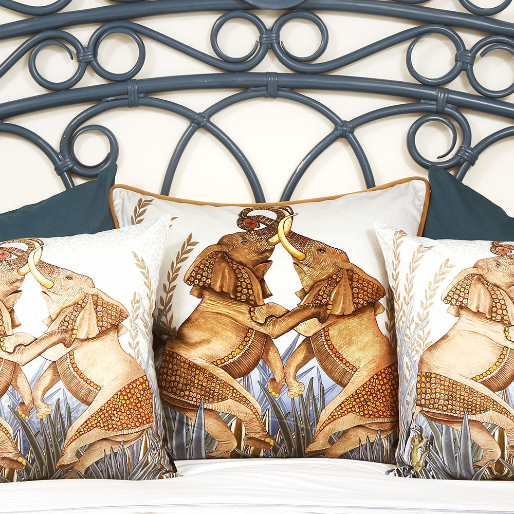 Lifestyle view of three elephant-printed throw pillows arrange on a bed in front of a dark blue wrought-iron headboard. 