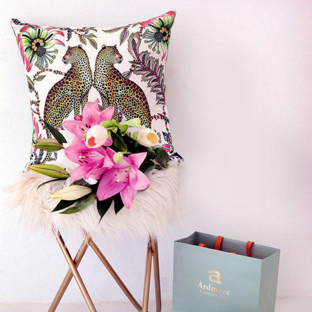 Lifestyle view of the white Lovebird Leopards Pillow displayed on a fur-trimmed stool with pink lilies positioned next to an Ardmore gift bag.