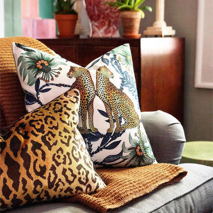 Lifestyle view of the Lovebird Leopards Pillow styled on a sofa next to a brown leopard-print cushion and a knitted throw blanket. 