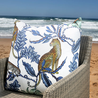 Lifestyle view of the blue Monkey Bean Pillow styled on an outdoor wicker armchair set against a beach and ocean backdrop. 