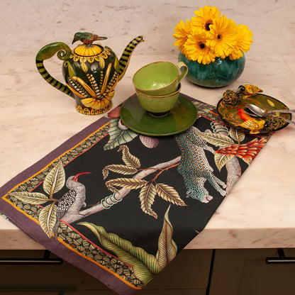 Pangolin Park Tea Towel styled on a kitchen counter with a green teacup and saucer, decorative teapot, spoon rest, and yellow flowers.